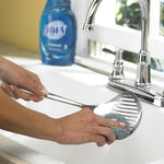 Hand using a Dawn steel mesh scrubber to clean a metal slotted spoon at a kitchen sink with Dawn bottle in background