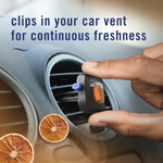 Hand placing a Febreze AUTO Bourbon & Orange vent clip into a circular car vent with dried orange slice accents