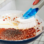 Hand-held Dish Daddy brush with a translucent blue handle attachment scrubbing heavy residue from a skillet surface.