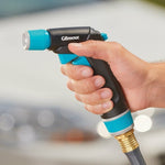 Hand holding a Gilmour hose nozzle, showing thumb on trigger and teal textured connector with hose fitting
