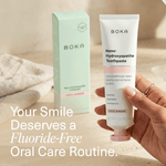 Hand holding a BOKA Nano-Hydroxyapatite toothpaste tube with its green box visible in soft natural light and text overlay about fluoride-free routine