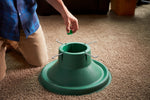 Person kneeling and holding a small green cap above a green plastic Christmas tree stand on carpet.