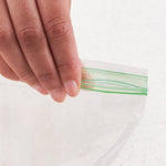 Close-up of fingers holding the green double-zipper closure of a clear sandwich bag against a light background