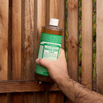 Hand holding a Dr. Bronner's Magic Soap almond bottle against a wooden fence background.
