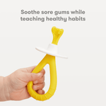 Hand holding a yellow Frida Baby silicone training toothbrush with white safety stopper against a light background