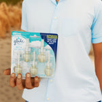 Person holding a blister pack of Glade PlugIns scented oil refills in hand, beach-like blurred background and teal packaging visible