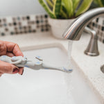 Hand holding a gray otter toothbrush under running faucet over a sink