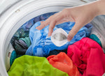 Hand holding a single mesh lint-and-hair remover ball over colorful clothing inside a washing machine drum.