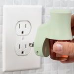 Hand holding a soft sea glass Mainstays plug near a wall outlet, showing bottle and prongs