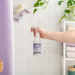 Hand holding a Mrs. Meyer’s probiotic daily shower spray bottle in a tiled bathroom scene with towels and plants