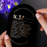 Hand holding a black card labeled 'N.37' with descriptive copy, purple flower petal visible at the edge.