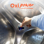 Hand holding a blue laundry detergent pac over a stainless steel washing machine drum with 'Oxi power' text