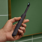 Hand holding a dark quip sonic electric toothbrush in front of green tiled background, angled view