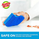 Hand holding a blue Scotch-Brite non-scratch sponge wiping a countertop with a small food smear nearby