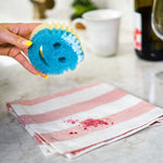 Hand holding a blue-and-yellow Scrub Mommy style sponge over a striped cloth with red stain on a countertop