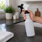 Hand holding a translucent spray bottle with black-and-white trigger over a kitchen counter