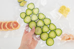 Hand holding a vacuum-sealed textured bag of cucumber slices with other sealed items blurred behind