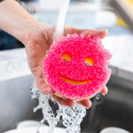 Hand holding a wet bright pink smile-faced scrub sponge under running faucet water over a stainless sink