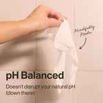 Hand holding a single white wipe in front of tiled wall with copy noting pH balanced and a small handwritten-style note.
