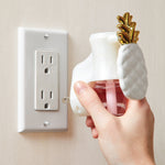 Hand inserting a white pineapple-shaped plug-in fragrance diffuser into a wall outlet with the pink refill bottle attached.