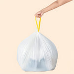 Hand holding the yellow drawstring of a full white trash bag lifted against a neutral beige background