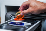 A hand placing an orange-and-clear laundry pod into a washing machine detergent drawer in a lifestyle product photo