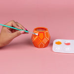 Hand painting an orange geometric ceramic planter with a brush and paint palette on a pink background