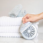 Hand peeling the top label off a round Odor Genie placed beside stacked white towels on a countertop
