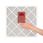 Hand placing a red Yankee Candle whole-home air freshener onto a square HVAC air filter on white background