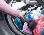 Hand placing a blue Downy Ball into a front-opening washing machine drum filled with colorful laundry.