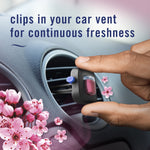 Hand placing a Febreze Auto vent clip into a car vent with cherry blossom graphics and text reading 'clips in your car vent for continuous freshness'.