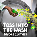 Hand pouring small green scent beads from a green measuring cap into the drum of a front-load washing machine with floral accents