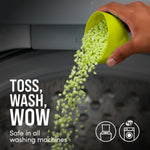Hand pouring small green scent booster beads from a scoop into a washing machine with caption toss, wash, wow