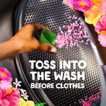 Hand pouring pink scent beads from a green measuring cap into a washing machine drum with floral accents