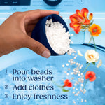 Hand pouring small white scent booster beads from a navy measuring cup toward a washing machine with instructional text
