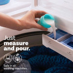 Hand pouring liquid fabric softener from a blue measuring cup into a washing machine dispenser drawer