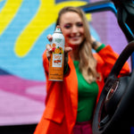Close-up of a woman offering a Citrus Magic Tropical Citrus car spray can from inside a car with a mural backdrop