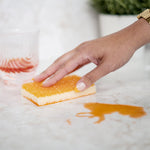 Hand pressing an orange-topped sponge on a countertop beside a spilled orange liquid demonstrating cleaning