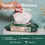 Hand pulling a wipe from a green Honest flushable wipes pack against a tiled background with headline text about gentle cleaning.