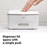 Hand pushing the lid of a white OXO wipe dispenser on tiled bathroom surface