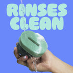 Hand rinsing the green Brite scrubber under running water with foam, blue background and large “RINSES CLEAN” text