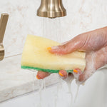 Hand holding a Scrub Daddy sponge under running faucet with soap suds rinsing from the sponge