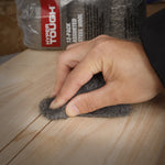 Hand holding a gray steel wool pad and rubbing a wooden surface with Hyper Tough package visible in background