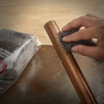 Hand scrubbing a copper pipe with a clump of gray steel wool with packaged product blurred in background