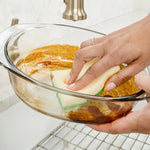 Hand scrubbing a soiled glass mixing bowl with a dual-sided sponge under a kitchen faucet