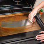 Hand scrubbing oven door interior with a Scrub Daddy sponge and Cif bottle visible at the edge