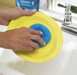 Hand using a round blue mesh scrubber to clean a yellow plate over a kitchen sink with faucet visible