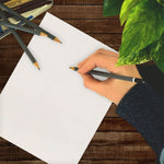 Top-down scene of a hand sketching on a blank sheet of white paper with pencils and a green plant on a wood surface