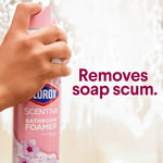 Hand pressing the top of a pink Clorox Scentiva Bathroom Foamer can next to text 'Removes soap scum.'