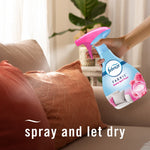 Hand holding a Febreze FABRIC spray bottle aimed at an orange couch pillow with text 'spray and let dry'.